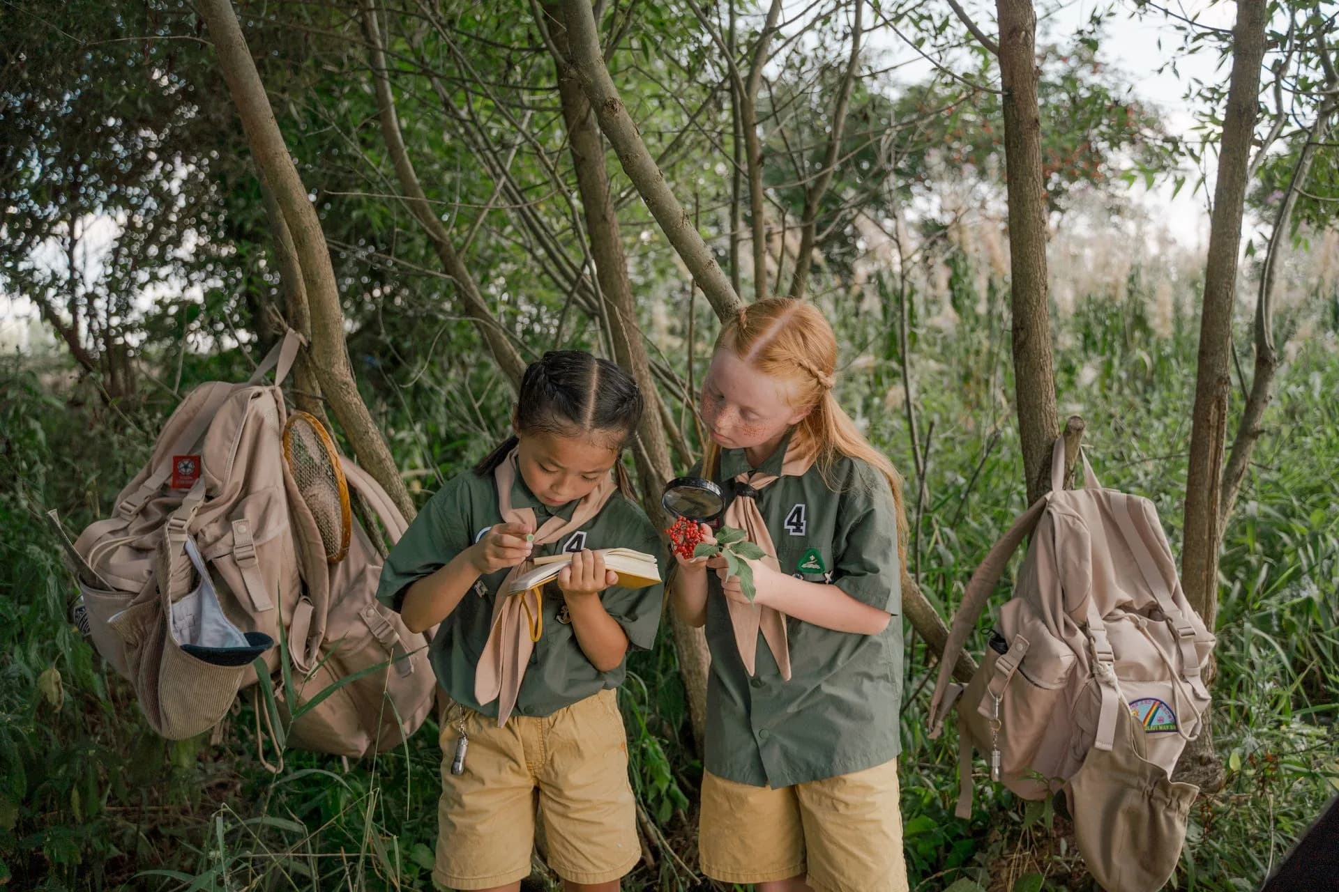 Two young students examining plants with a magnifying glass and notebook during an outdoor woodland field study